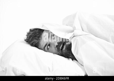 Man with beard and mustache hiding in bed under blanket at home. Guy with surprised face lay under white blanket. Stock Photo