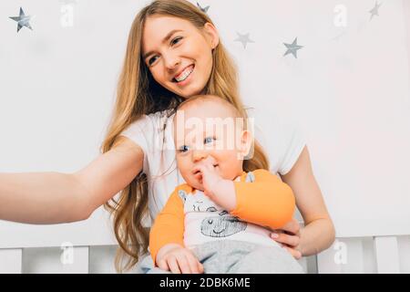 A happy mother and a small baby relax while sitting on the bed and take a selfie on their mobile phone. Stock Photo