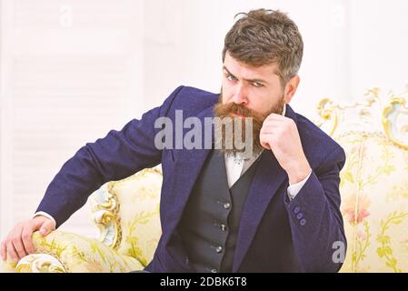 Elite lifestyle concept. Man with beard and mustache wearing classic suit, sits on old fashioned armchair or sofa. Macho attractive and elegant on serious face and thoughtful expression. Stock Photo