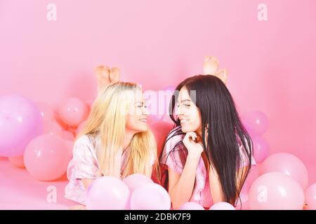 Girls lay on belly near balloons, pink background. Friendship concept. Sisters or best friends in pajamas at girlish pajamas party. Blonde and brunette on smiling faces have fun at domestic party. Stock Photo