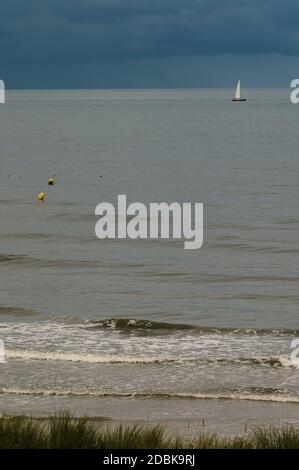 In the calm and gray sea there are yellow buoys and a sailing boat under a sky with threatening clouds. Stock Photo