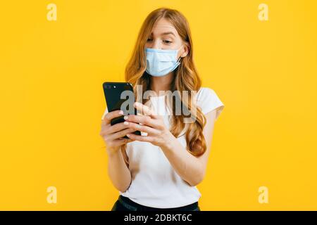 A young woman, wearing a protective medical mask, uses a mobile phone on a yellow background. The concept of quarantine, coronavirus Stock Photo