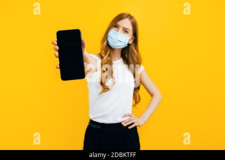 A young woman in a protective medical mask shows a blank smartphone screen to copy space on a yellow background. Concept of quarantine, coronavirus Stock Photo