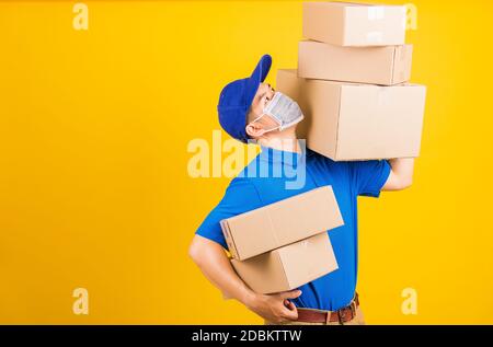 Asian young delivery worker man in blue t-shirt and cap uniform wearing face mask protective lifting stack a lot of boxes, under coronavirus or COVID- Stock Photo