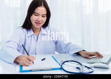 Asian doctor young beautiful woman smiling using working with PC desktop computer and her writing something on paperwork or clipboard white paper at h Stock Photo