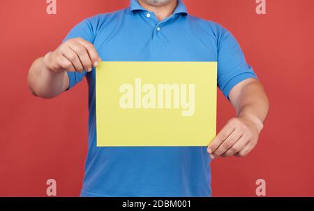 man in blue clothes holds an empty sheet of yellow paper on a red background, place for inscription Stock Photo