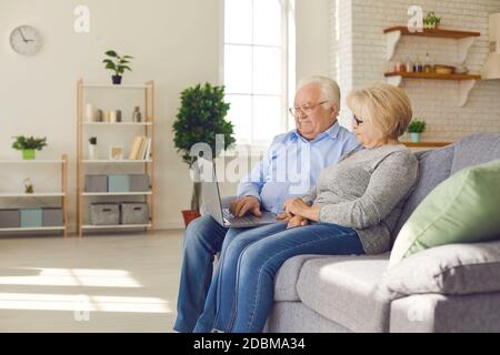 Happy cute senior couple sitting on couch at home and using laptop computer together Stock Photo