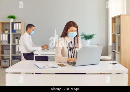 Female office worker in medical mask working with laptop and keeping distance with her colleague. Stock Photo