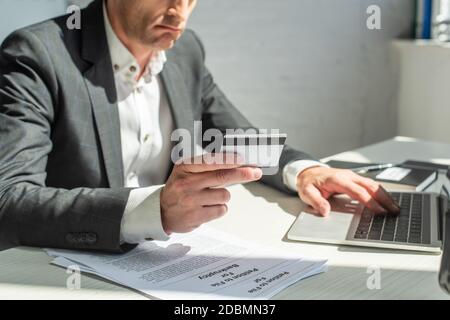 Cropped view of sad businessman with credit card, typing on laptop, while sitting at workplace on blurred background Stock Photo