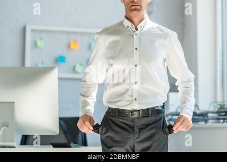Cropped view of businessman showing empty pockets, while standing near workplace on blurred background Stock Photo