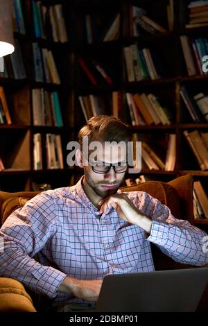 Serious young business man using laptop working late online sitting in chair. Stock Photo