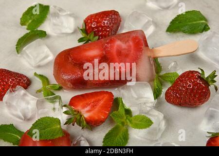 Close up fruit ice cream popsicle with fresh strawberry, green mint leaves and ice cubes on white table surface, elevated top view, directly above Stock Photo