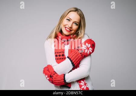 Lovely girl in mittens embracing herself. blonde woman in red knitted scarf and mittens embracing herself and smiling at camera. Stock Photo