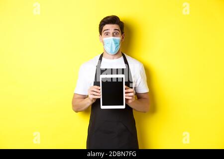 Concept of covid-19, small business and pandemic. Surprised waiter in black apron and medical mask showing tablet screen, standing over yellow Stock Photo