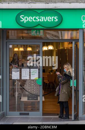 a young woman standing in the doorway of a specsavers store in a town centre looking at her mobile phone waiting for her appointment. Stock Photo