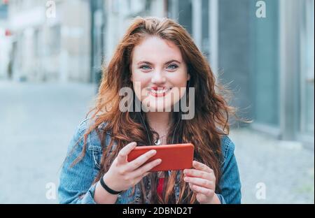 Portrait of smiling red curled long hair caucasian teen girl walking on the street and video chatting using the modern smartphone. Modern people with Stock Photo