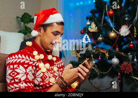 Man using smartphone for Christmas. Guy celebrating New year alone at home wearing Santa's hat, lights by Christmas tree. Stock Photo
