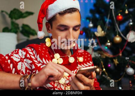 Man using smartphone for Christmas. Guy celebrating New year alone at home wearing Santa's hat, lights by Christmas tree. Stock Photo