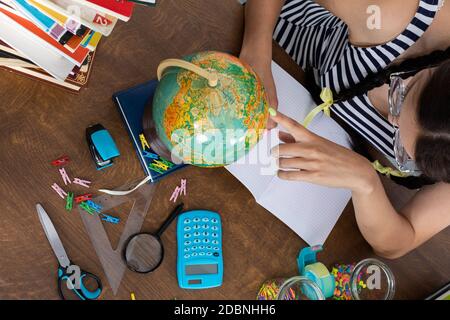 A small globe is standing on the bench. The schoolgirl is sitting at the school desk. School supplies scattered across the school bench. Stock Photo