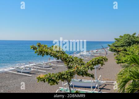 Black volcanic sand beach from nearby Agung volcano. Tropical paradise with palm trees and majestic volcano in the background. View from above. Stock Photo