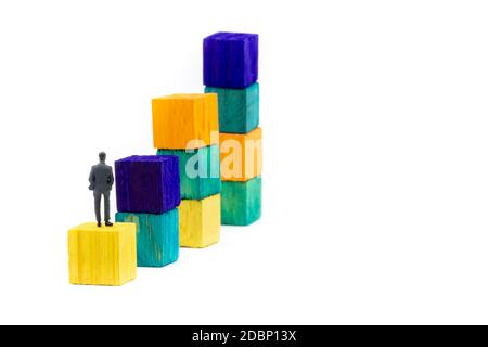 Miniature figurine posed as businessman sitting alone on a wooden cube looking up to colourful stacks as business chart, minimalist abstract concept i Stock Photo