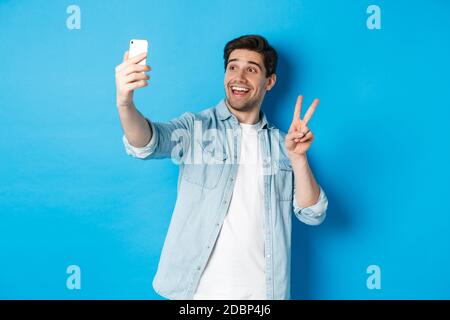 Happy man taking selfie and showing peace sign on blue background, holding mobile phone Stock Photo