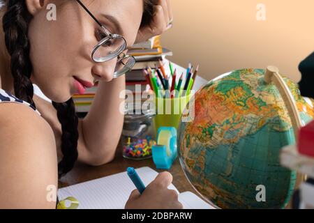 A small globe is standing on the bench. The schoolgirl is sitting at the school desk. School supplies scattered across the school bench. Stock Photo