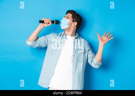 Handsome guy in medical mask singing in microphone, standing over blue background Stock Photo