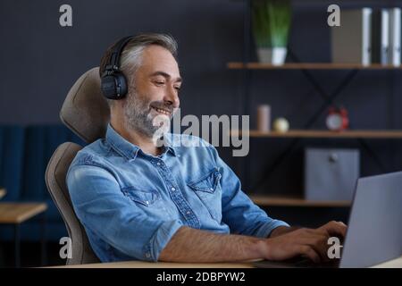 Portrait of handsome grey-haired man wearing headphones sitting at workplace. Senior male freelancer working in home office with laptop. Studying Stock Photo