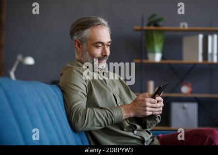 Smiling grey-haired man using mobile phone apps, texting message, browsing internet, shopping online. Looking at smartphone and smiling, sitting at Stock Photo
