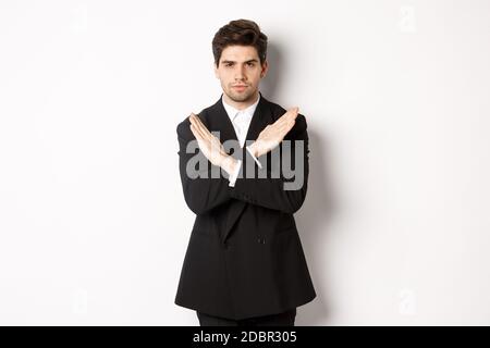Portrait of serious and concerned man in black suit, showing stop gesture and frowning, making cross to prohibit or decline something, standing over Stock Photo
