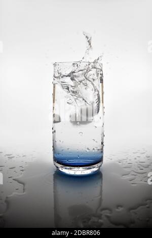 An ice cube dropping into a glass of water creating a splash. A reflection of the glass in the pool of water around the drink. Stock Photo