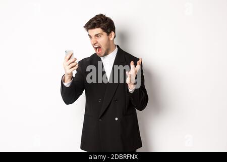 Portrait of angry businessman in black suit yelling at mobile phone, having an argument on video call, standing mad over white background Stock Photo