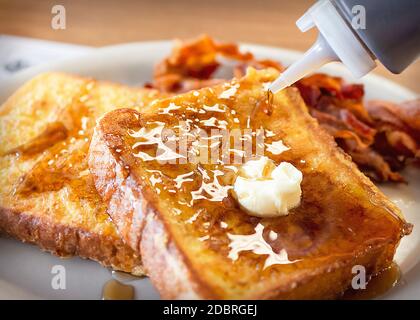 Two pieces of French Toast on a white plate with bacon blurred in the background.  Daub of butter melts ontop while maple syrup is being poured.  Phot Stock Photo