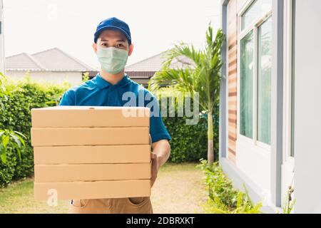 Asian young delivery man courier sending and holding fast food pizza boxed in uniform he protective face mask service customer at home door, under cur Stock Photo