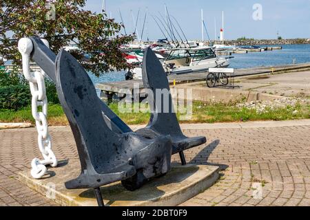 A large black anchor with a white chain stands on a concrete slab against a bay with moorings for boats Stock Photo