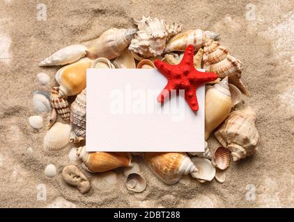 Blank white leaf for text or greetings lies on seashells. Seashells on the sand, marine theme. Top view. Stock Photo