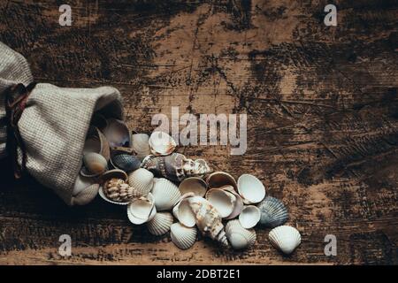 Linen bag with small seashells on old wooden background. Place for text. Retro toned. Old burnt wood texture board. Stock Photo