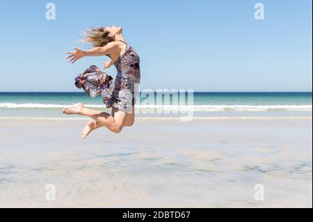 Middle-aged blonde woman in a green dress jumping with her arms open on the beach Stock Photo