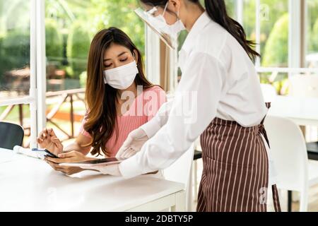 Asian waitress wear facemask and face shield using tablet to show restaurant electronic menu and recommend to customer. New normal technology and life Stock Photo