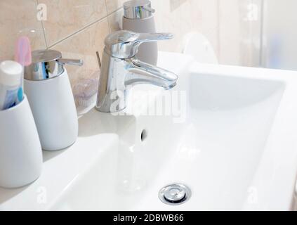 a white ceramic sink with a chromed tap with soap and toothbrush containers in a bathroom. Interior of a house. Stock Photo