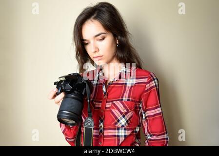 Tired serious woman photographer in red shirt holding camera and looking at bad,failed and unsuccessful photos on beige background. Stock Photo