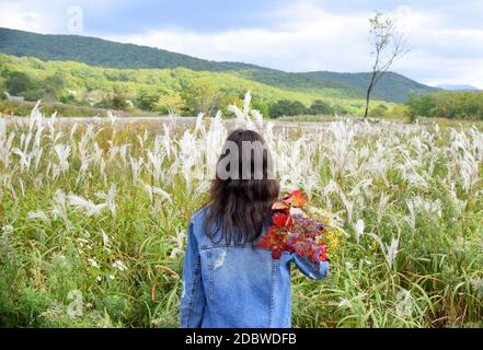 Young woman holding bouquet of autumn wildflowers and branches with red leaves on her shoulder, standing with her back to the camera Stock Photo