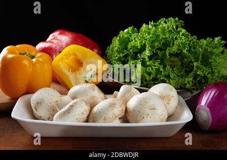 raw mushrooms in plastic wrap, red and yellow peppers, green lettuce and purple onions on a brown wooden table, ingredients for cooking Stock Photo