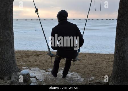 Adult man in a coat swings on a swing and looks thoughtfully at the sea on a cold evening Stock Photo