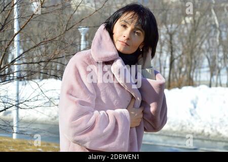 Forty five year old Russian woman with bright brown eyes, in pink coat smiling, looking at the camera on an early spring  day in city Stock Photo