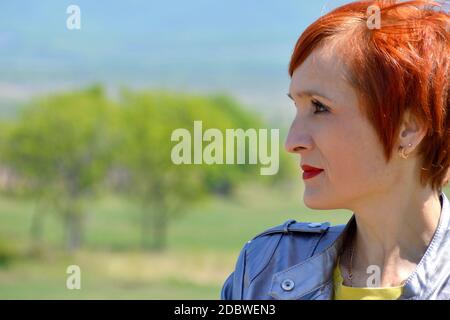 Portrait of Russian confident woman with short red hair, looking left from right side on a background of green field with trees. Stock Photo