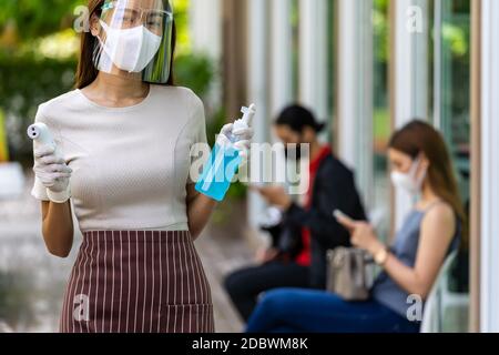 Portrait attractive asian waitress wear face mask and shield hold thermometer and alcohol gel sanitizer with background of social distancing customer Stock Photo