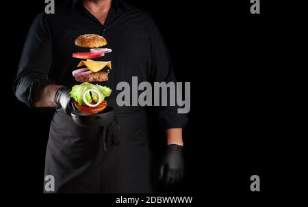 adult man in a black uniform holding a cast iron round frying pan with frozen cheeseburger ingredients: sesame bun, cheese, tomato, onion, meat cutlet Stock Photo