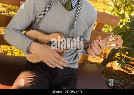 Young man sitting on bench in the park and playing ukulele. Stock Photo